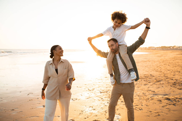 Glückliche dreiköpfige multiethnische Familie, die abends am Meer spazieren geht, Vater trägt Sohn auf Schultern, während sie gemeinsam draußen spazieren gehen und den warmen Sommertag genießen - Foto, Bild