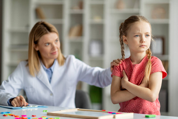 Psychoterapeutka pocieszająca zdenerwowaną dziewczynkę podczas sesji terapeutycznej w biurze, specjalistka od rozwoju dziecka próbująca uspokoić pacjentkę, obrażone dziecko stojące ze złożonymi ramionami - Zdjęcie, obraz