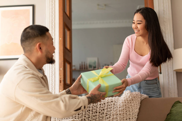 Dárek pro manžela na Valentýna, narozeniny a výročí. Usmívající se asijka dává dárek zabalený do krabice pro muže v obývacím pokoji interiéru, selektivní zaměření. Koncept rodinné dovolené - Fotografie, Obrázek