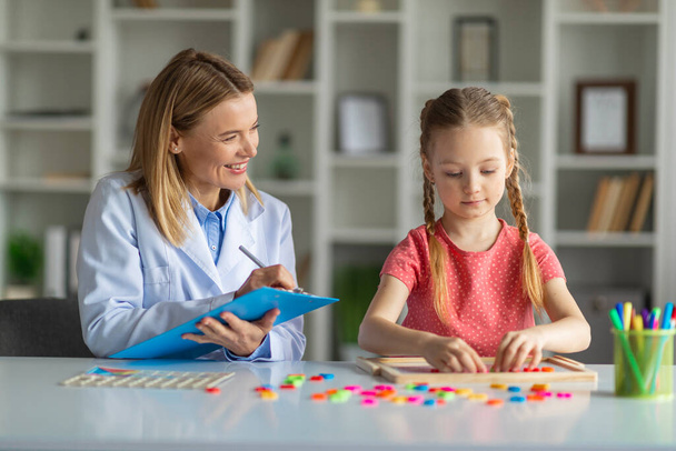 Kids Psychology Concept. Smiling Psychotherapist Lady Taking Notes During Session With Little Girl, Cute Female Child Making Word With Colorful Letters On Table, Enjoying Development Games, Closeup - Fotografie, Obrázek