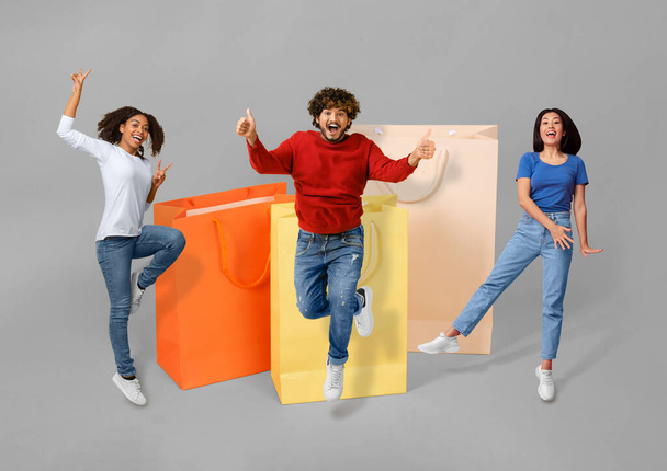Happy positive multiethnic people shopaholics millennial women and man wearing casual outfit posing by huge colorful shopping bags, jumping and gesturing, isolated on background, collage - Photo, Image