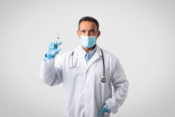 Серйозно впевнений кавказький зрілий лікар у білому пальто, захисна маска і рукавички тримають шприц, ізольовані на сірому фоні, студія. Вакцинація та лікування захворювань, охорона здоров'я, медицина
 - Фото, зображення