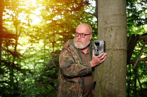 Ο Χάντερ βάζει μια κάμερα σ 'ένα δέντρο στο δάσος. Οι κάμερες Trail χρησιμοποιούνται συχνά από κυνηγούς για αυτόματη φωτογράφηση ή βιντεοσκόπηση της άγριας ζωής στο δάσος. - Φωτογραφία, εικόνα