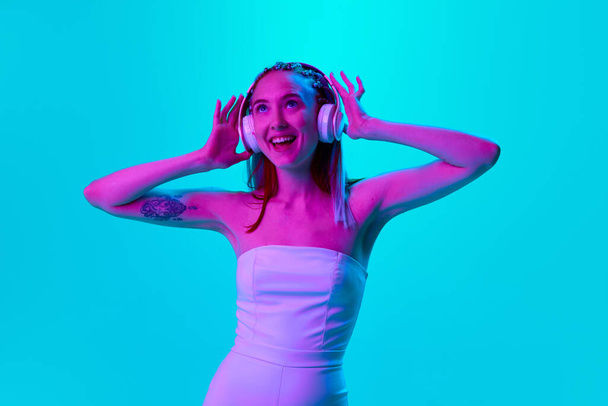Portrait of beautiful young woman listening to music in headphones and smiling against blue studio background in neon light. Concept of youth, emotions, lifestyle, beauty, lifestyle, ad - Photo, Image
