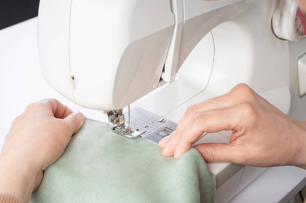 Female hands stitching white fabric on modern sewing machine at workplace in atelier. Women's hands sew pieces of fabric on a sewing machine closeup. Handmade, hobby, small business concept - Photo, Image