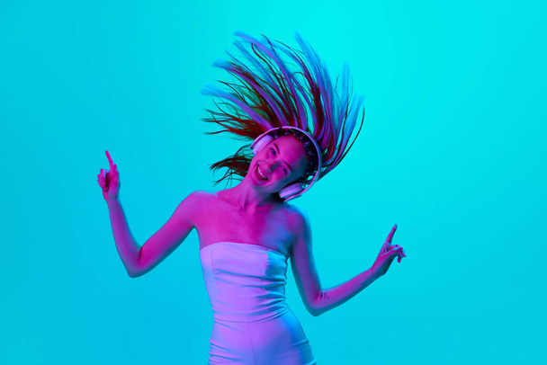 Energy and fun. Portrait of beautiful young woman listening to music in headphones and dancing against blue studio background in neon light. Concept of youth, emotions, beauty, lifestyle, ad - Фото, изображение