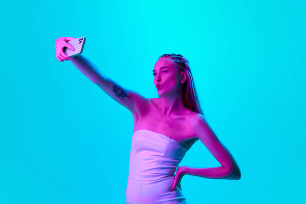 Stylish, beautiful young girl taking selfie with mobile phone against blue studio background in neon light. Social media influencer and communication. Concept of youth, emotions, beauty, lifestyle, ad - Foto, imagen