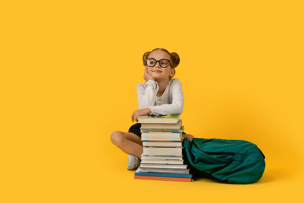 Cute Elementary School Girl Sitting At Books Stack Over Yelow Background, Smiling Dreamy Preteen Female Child With Backpack Posing In Studio, Happy Pupil Kid Looking Aside At Copy Space - 写真・画像