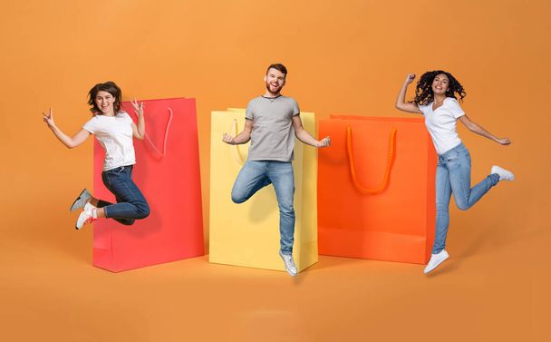 Big sale, discount, black friday deal. Excited cheerful multiethnic millennials happy man and two ladies jumping by huge colorful shopping bags, celebrating season sale, colorful background, collage - Photo, Image