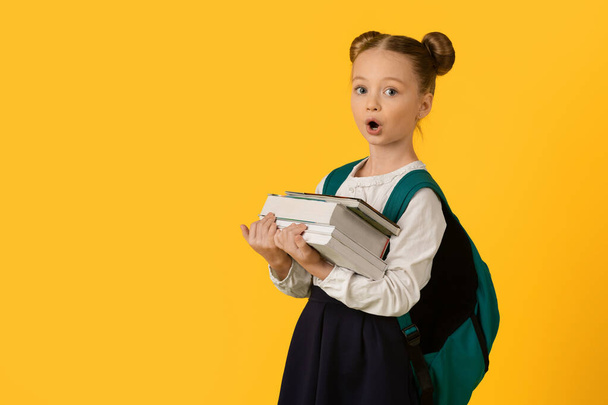 Πίσω στο σχολείο. Πορτρέτο του έκπληκτος Nerdy Schoolgirl μεταφέρουν βαριά βιβλία και κοιτάζοντας κάμερα, χαριτωμένο Preteen θηλυκό παιδί κουρασμένος από την εργασία στο σπίτι, στέκεται απομονωμένο σε κίτρινο φόντο, αντίγραφο χώρο - Φωτογραφία, εικόνα