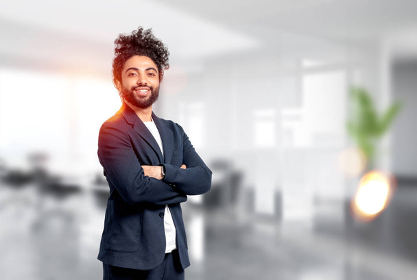 Smiling handsome businessman wearing formal wear is standing in crossed arms pose at blurred office workplace in background. Concept of model, successful business person, confidence - Fotó, kép