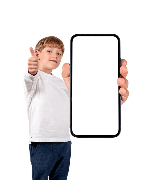 Niño sonriendo, pulgar hacia arriba y sosteniendo el teléfono inteligente con gran pantalla de espacio de copia simulada, aislado sobre fondo blanco. Concepto de aplicación móvil - Foto, imagen