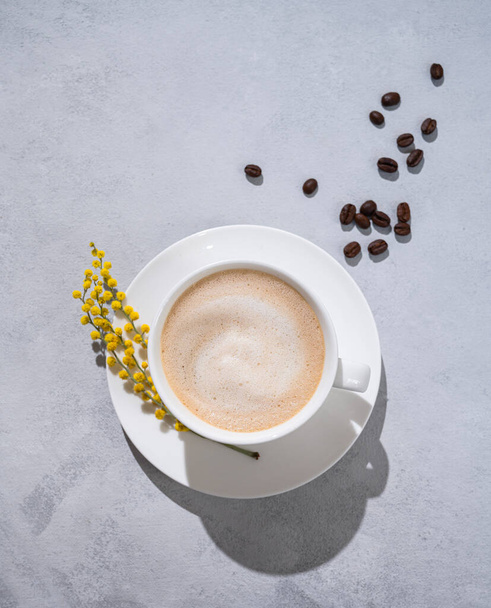 Una taza de café capuchino con mimosa flor de primavera sobre un fondo claro con granos de café. Concepto del 8 de marzo, feliz día de la mujer. Vista superior y espacio para texto - Foto, Imagen