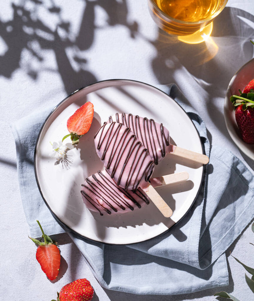 Gâteau au chocolat éclate crème glacée sur un bâton en glaçure rose sur une assiette avec des baies sur un fond clair avec une tasse de thé et d'ombre. Bonbons maison sains. De la nourriture. Vue du dessus. - Photo, image
