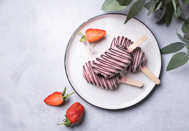 Chocolate cake pops ice cream on a stick in pink glaze on a plate with berries and eucaluptus branch on a light background. Healthy homemade sweets. Party food. Top view. - Фото, изображение