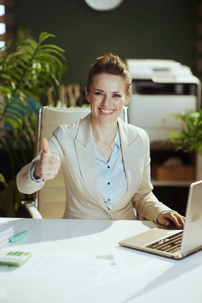 Portrait of smiling modern woman worker in a light business suit in modern green office with laptop showing thumbs up. - Zdjęcie, obraz