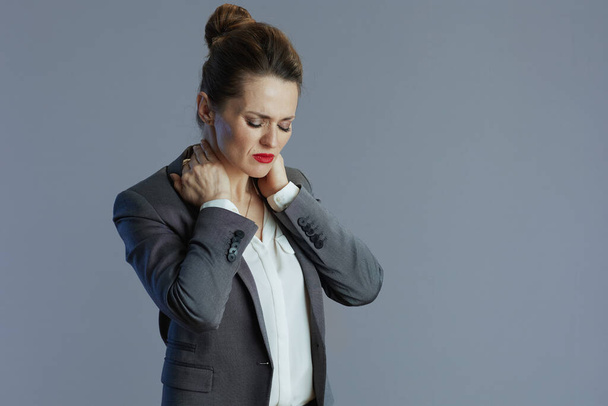 stressed stylish 40 years old business woman in gray suit having neck pain against gray background. - 写真・画像