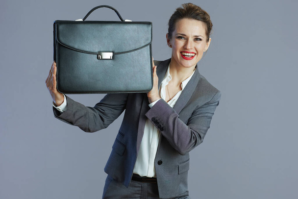 happy modern 40 years old woman worker in gray suit with briefcase against gray background. - 写真・画像