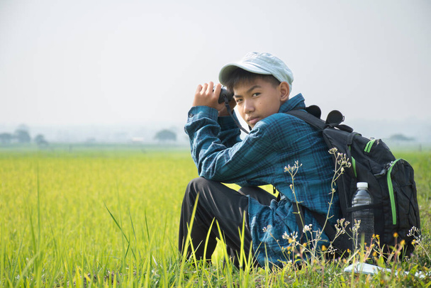 Asian boy in plaid shirt and cap sitting on paddy field in the morning and holding binoculars in  hands to watch birds which flying on the sky and eating fish in rice paddy field, summer vacation. - Zdjęcie, obraz