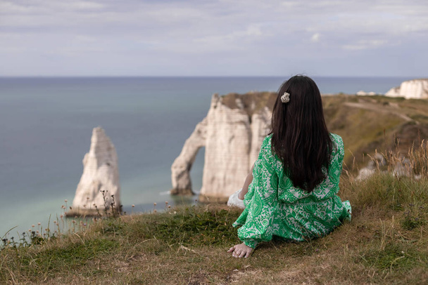 髪の毛が流れる緑のドレスを着た若い白人のブルネットの女の子が、エチケットの暗い春の日に北の海と自然のアーチの景色を楽しむ山の上の後ろから座っています - 写真・画像