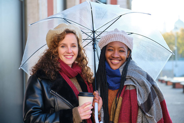 Two multiracial female friends looking at camera and smiling in winter, holding an umbrella and a coffee cup. - Foto, immagini