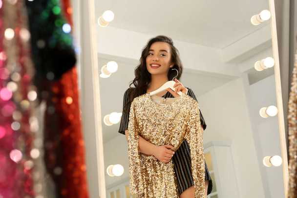 Beautiful young woman with golden sequin dress near mirror in boutique. Choosing party clothes - Zdjęcie, obraz
