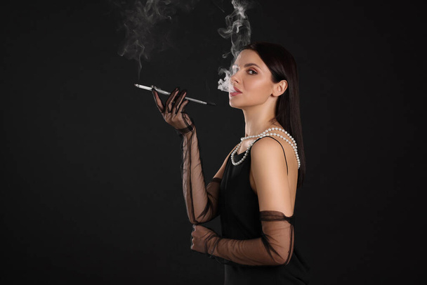 Woman using long cigarette holder for smoking on black background, space for text - Φωτογραφία, εικόνα