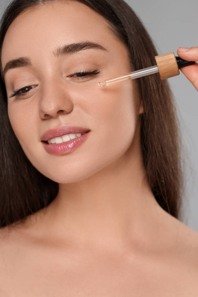 Beautiful young woman applying essential oil onto face on grey background, closeup - Фото, зображення