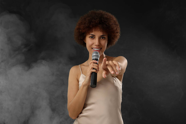 Curly young woman with microphone singing in smoke on black background - Foto, Imagen