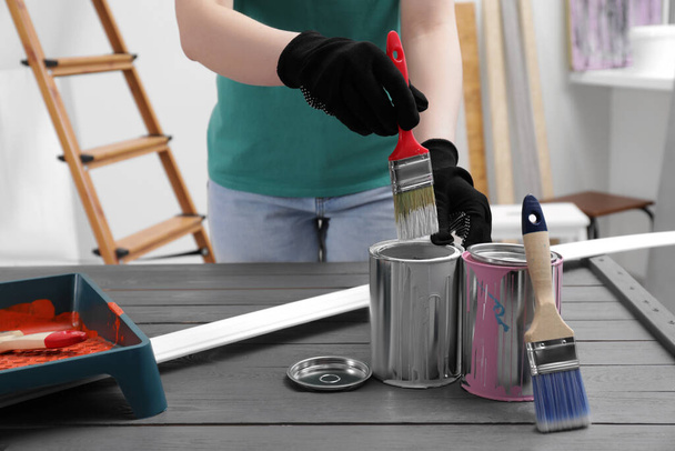 Woman dipping brush into can of white paint at black wooden table indoors, closeup - Foto, Bild