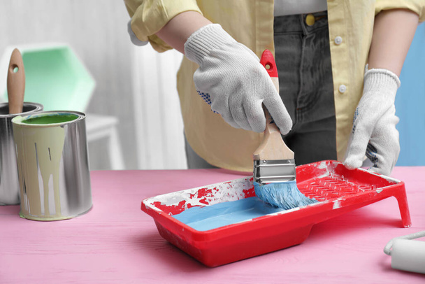 Woman taking light blue paint with brush from tray at pink wooden table indoors, closeup - Foto, imagen
