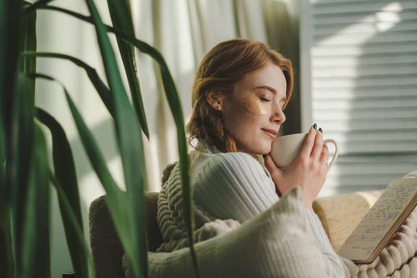 Pretty dreamy young woman sitting on couch covered with a blanket, drinking coffee indoors at home room. Cosy scene, cozy home concept - Fotografie, Obrázek