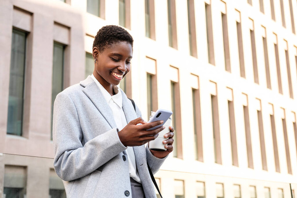 retrato horizontal de una elegante joven africana. Ella está de pie cerca del edificio de oficinas y usando su teléfono inteligente - Foto, imagen