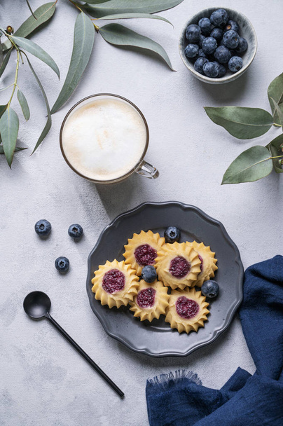 ダークプレートに自家製ベリークッキー、ユーカリの枝を背景にブルーベリーとカップにミルクフォームとカプチーノやラテコーヒー。コンセプト春の朝の朝食。トップ表示. - 写真・画像
