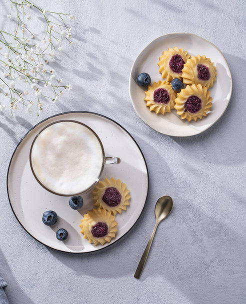 Cappuccino or latte with milk foam in a cup with homemade berry cookies and blueberries on a light background with gypsophila branches and shadow. Concept spring morning breakfast. Top view  - Fotó, kép