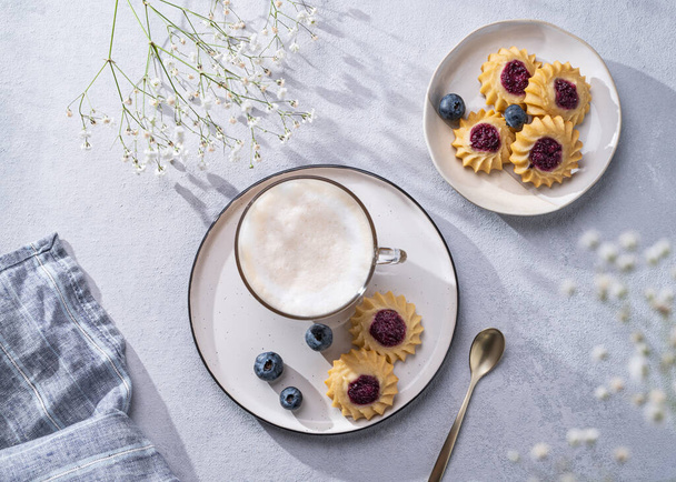Cappuccino or latte coffee with milk foam in a cup with handmade berry cookies and blueberries on a light background with gypsophila branches and shadow. Concept spring morning breakfast. Top view. - Fotó, kép
