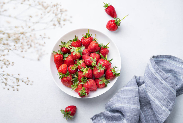 Juicy strawberries in a white bowl on a light blue background with napkin and flowers. Farm ripe healthy berries. Top view and copy space. - Photo, image