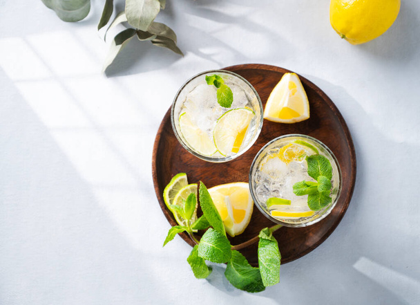 Lemonade drink with fresh lemon, lime, mint and ice. Refreshing citrus mojito cocktail on a light background with shadow. Summer cold drinks concept. Top view and copy space. - 写真・画像