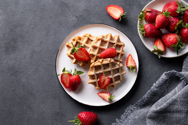 Homemade soft waffles with fresh strawberries in a plate on a dark background. Traditional Belgian waffles. Healthy breakfast. Top view and copy space. - Zdjęcie, obraz