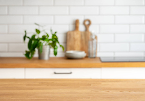 Holztischplatte mit freiem Platz für Produktmontage oder Mockup gegen verschwommene weiße Küche mit Schneidebrett und Pflanze im skandinavischen Stil im Morgenlicht - Foto, Bild
