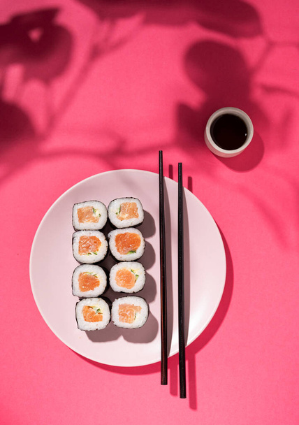 Japanse sushi roll met zalm, komkommer en sojasaus met eetstokjes in de plaat op roze ondergrond met hard schaduwbloemblaadje. Concept voedselfotografie. Bovenaanzicht en kopieerruimte. - Foto, afbeelding