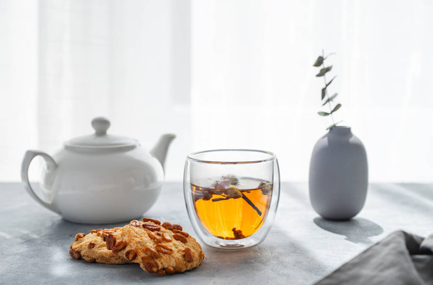 Un vaso de té de hierbas, tetera y galletas caseras en el fondo de una ventana con luz del sol de la mañana. Concepto de bebida para el desayuno. - Foto, imagen