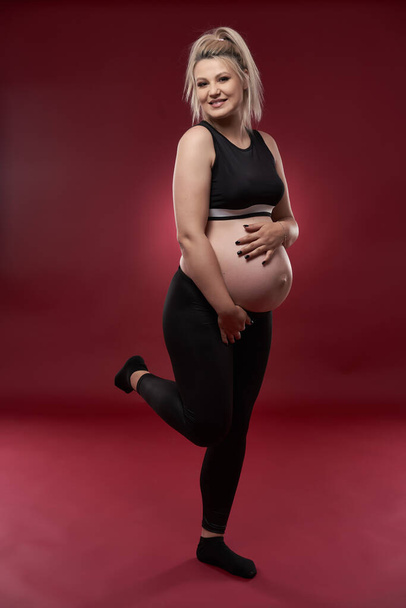 Beautiful pregnant young blonde woman in black clothes, studio shot on red background    - Foto, imagen