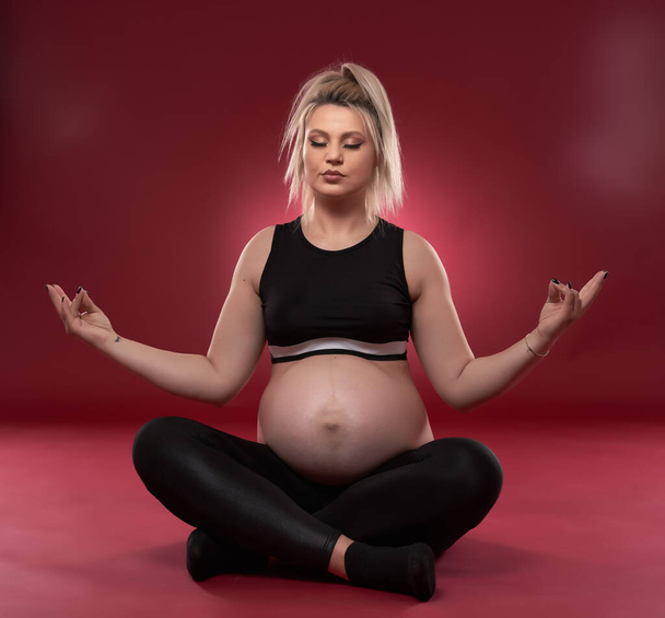 Beautiful pregnant young blonde woman in black clothes, studio shot on red background    - Fotó, kép