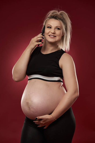 Pregnant woman speaking on mobile phone, studio shot on red background  - Foto, imagen