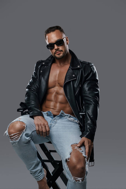Attractive male model with a muscular shirtless torso poses in ripped jeans, a black leather jacket, and dark sunglasses while seated on a studio chair against a grey backdrop - Photo, image