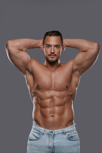Opvallend knap mannelijk model met een goed gebouwde lichaamsbouw poseert shirtloos in jeans tegen een grijze achtergrond, pronkt met zijn gebeitelde buikspieren en betoverende blik - Foto, afbeelding