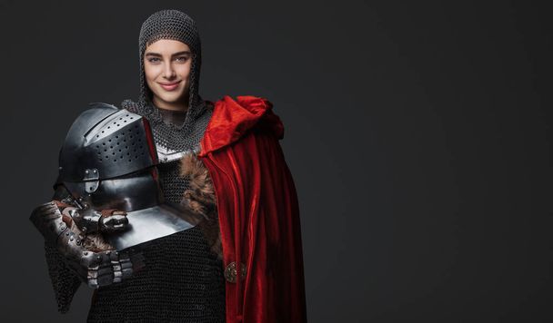 Selbstbewusste Ritterin in mittelalterlicher Rüstung, breit lächelnd und mit Helm in Pose, drapiert in einem roten Mantel, auf grauem Hintergrund - Foto, Bild