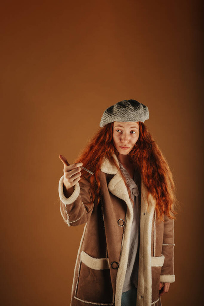 Ginger κορίτσι κοιτάζοντας μακριά, ενώ κρατώντας ένα τσιγάρο στο χέρι της και στέκεται στο στούντιο - Φωτογραφία, εικόνα