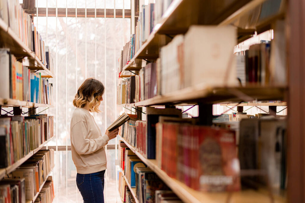 Kaunis tyttö lukee kirjaa seisoessaan kirjahyllyjen välissä - Valokuva, kuva